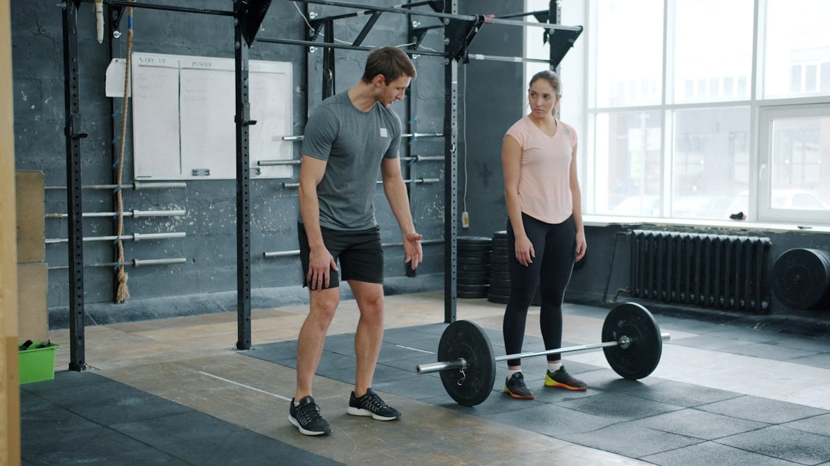 Male instructor is teaching young woman weighlifting techniques talking and gesturing in gym, girl is standing near heavy barbell looking at coach.